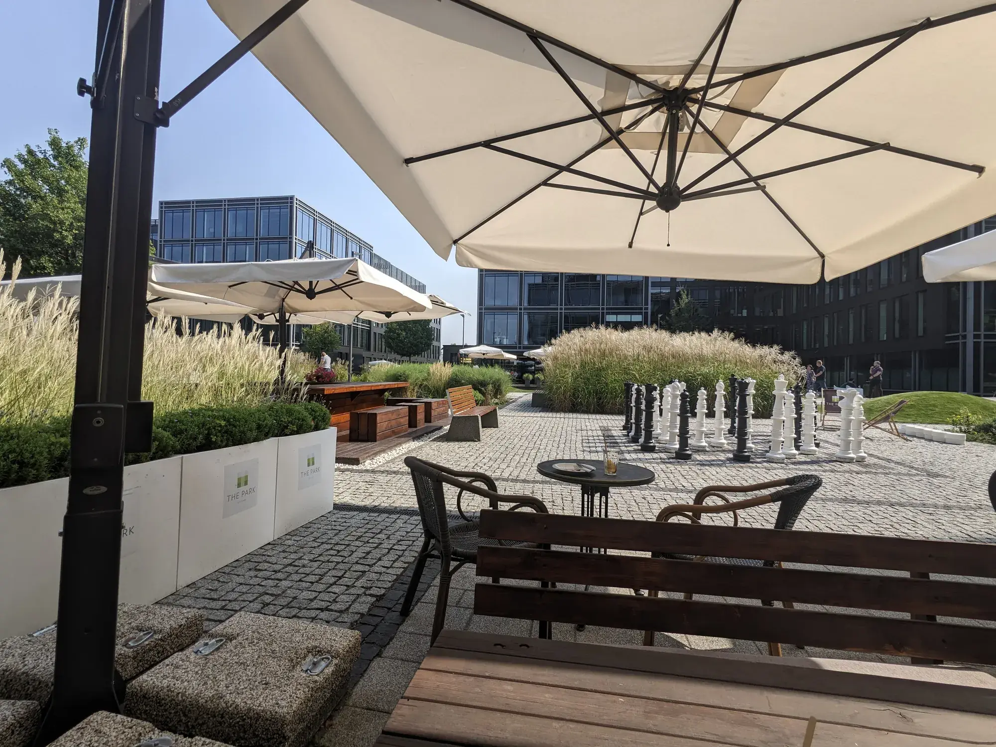 Patio The Park Warsaw se slunečníky nad lavicemi a stoly, v pozadí zahradní šachy