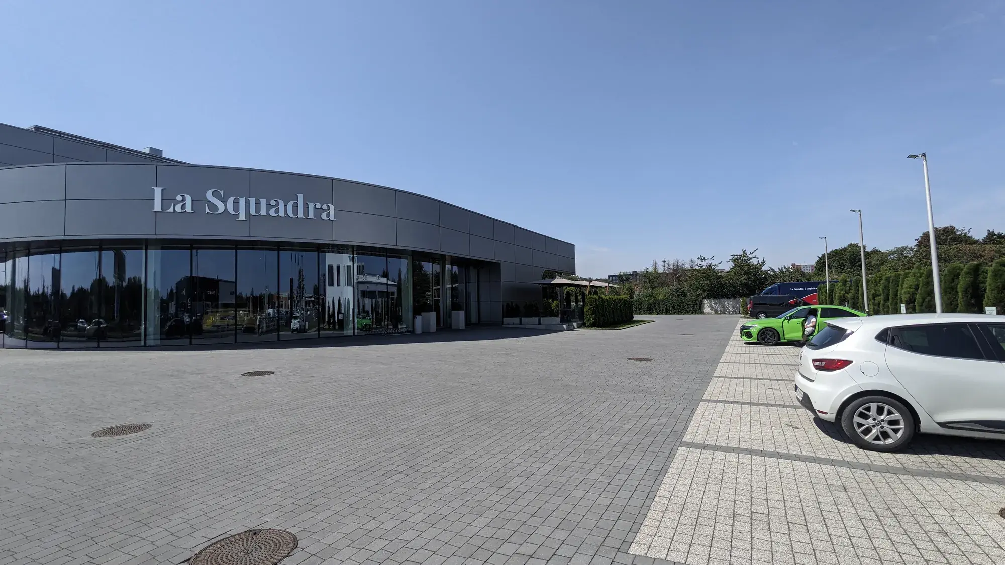 Budova La Squadra s logem na fasádě, v pozadí slunečníky nad terasou restaurace