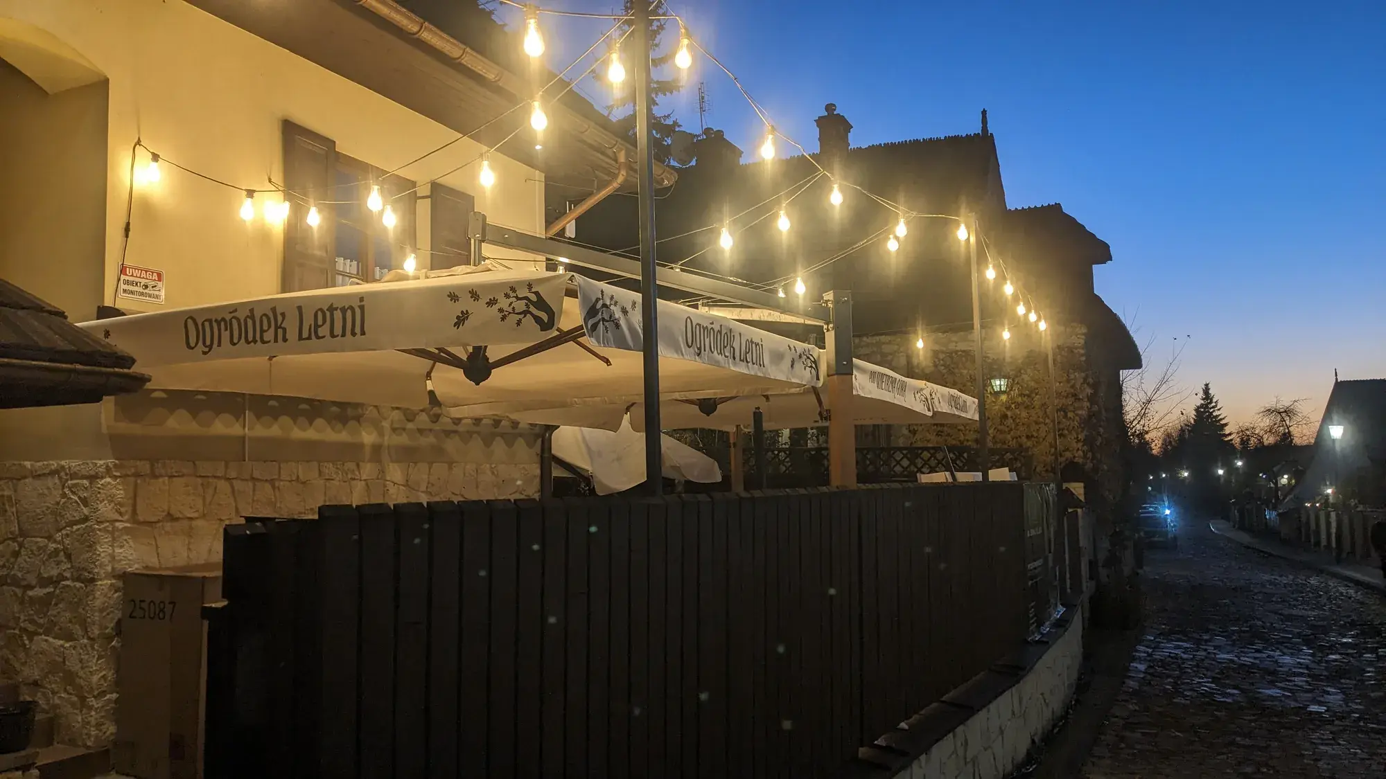Letní předzahrádka restaurace Pod Wietrzną Górą za soumraku, slunečníky s potiskem „Ogródek Letni“ pod girlandami žárovek