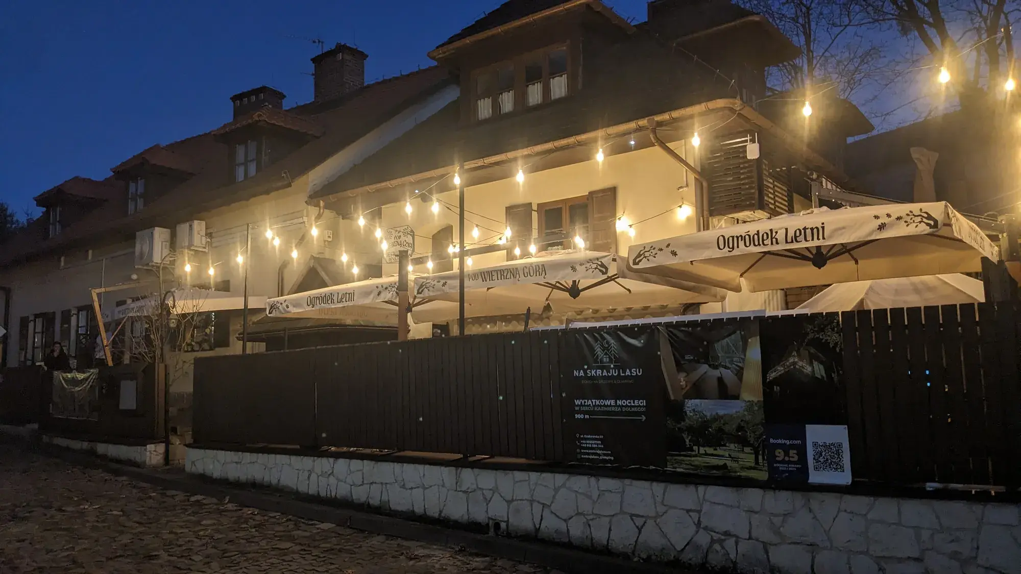 Restaurace Pod Wietrzną Górą v noci, slunečníky nad předzahrádkou za dřevěným plotem